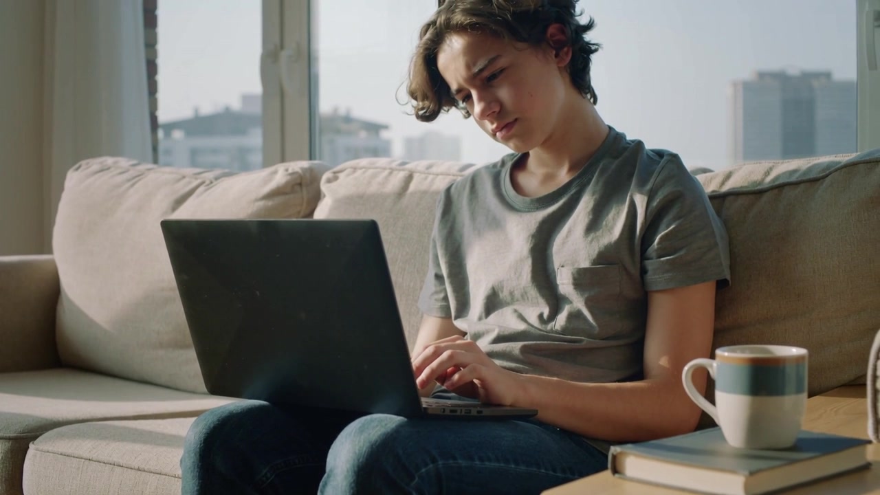 Typing teen male wearing jeans leaning forward, studying on sofa at home with laptop and mug