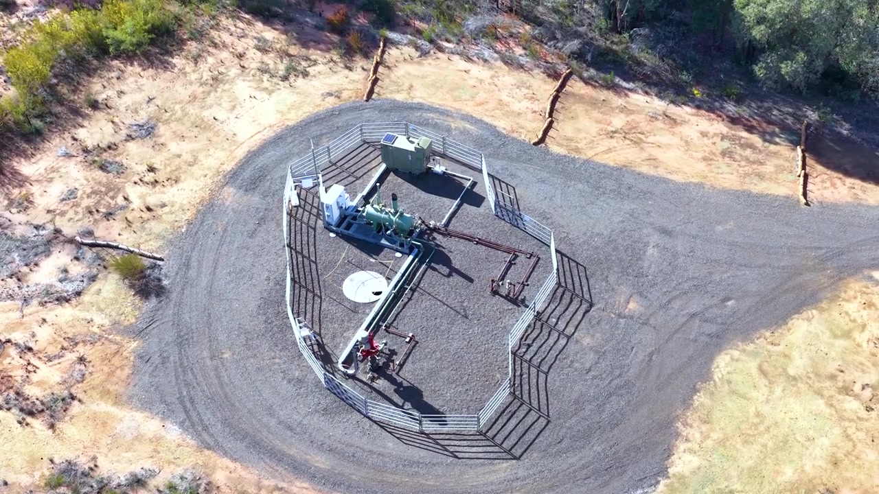Aerial view over a fenced industrial gas well pad located within remote Australian bushland.