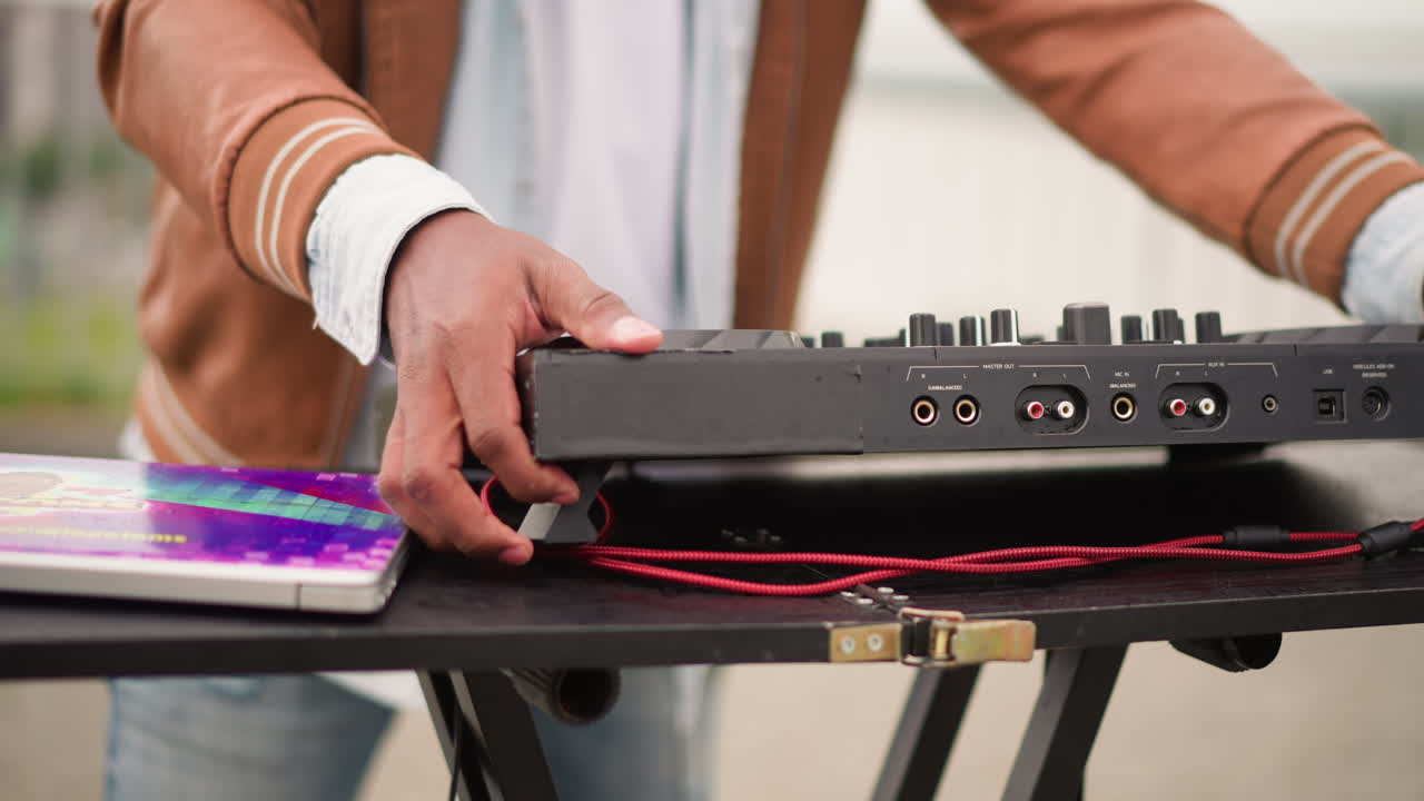 Configuração de DJ ao ar livre com controlador e tablet, mãos colocando controlador portátil em suporte dobrável ao lado de tablet colorido, cabo vermelho enrolado na capa, jaqueta casual e jeans, fundo urbano, nublado