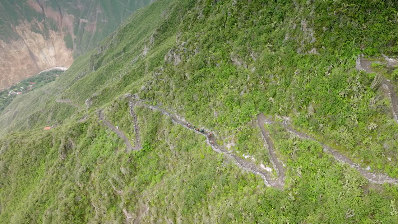 Drone shot in reverse, following hikers trekking down the path toward the Oasis of Sangalle, with the river flowing through the Colca Canyon in the background.