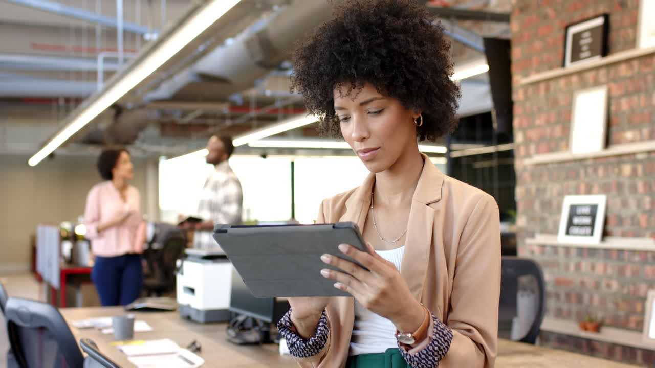 Biracial woman using tablet in modern office, open workspace with colleagues