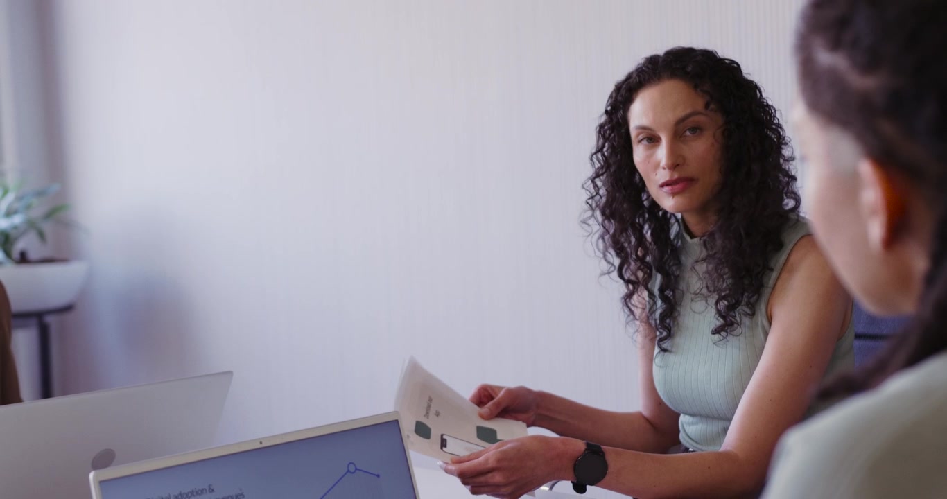 Donna afroamericana che tiene in mano dei fogli e discute di grafici a un tavolo riunioni mentre un collega indica il PC con una penna