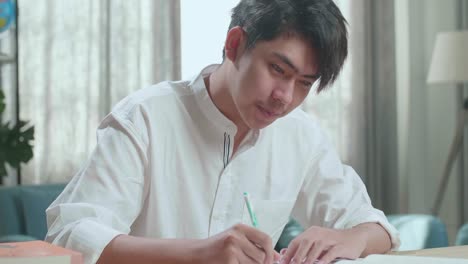 Close Up Of A Happy Asian Man Student Writing In Notebook On The Table While Studying At Home