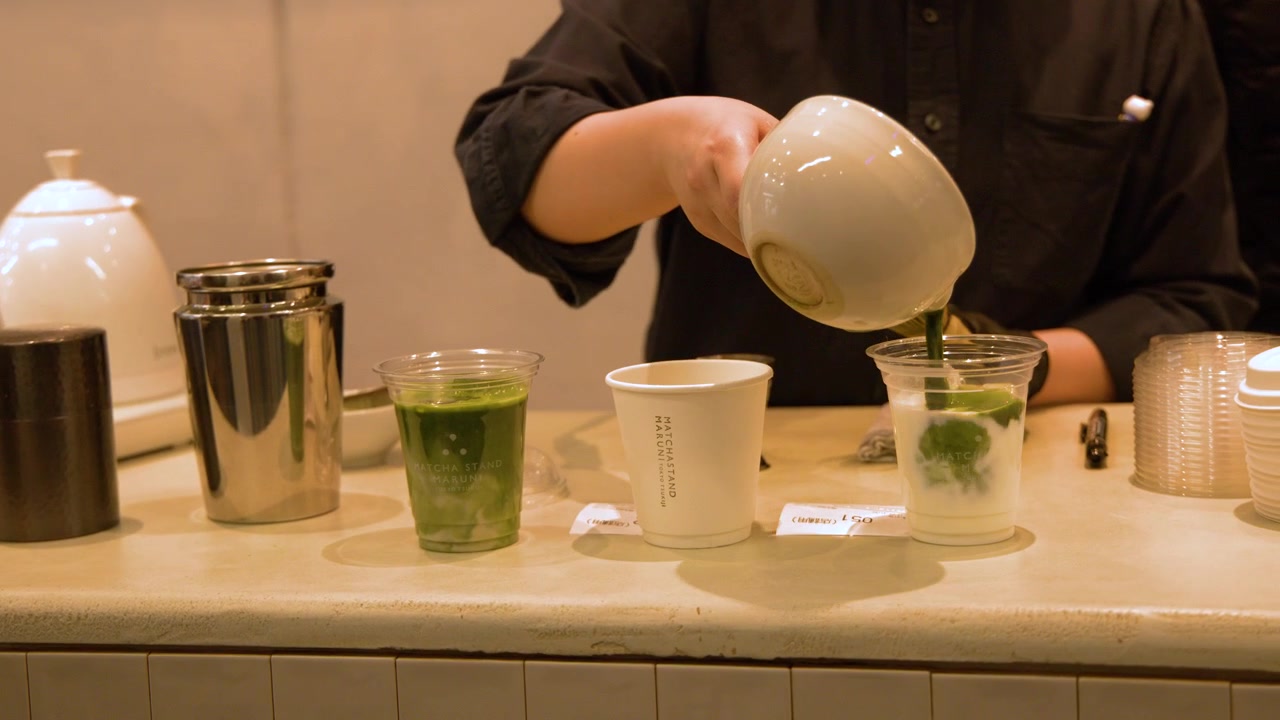 A barista pours concentrated green matcha tea from a ceramic bowl into takeaway cups.