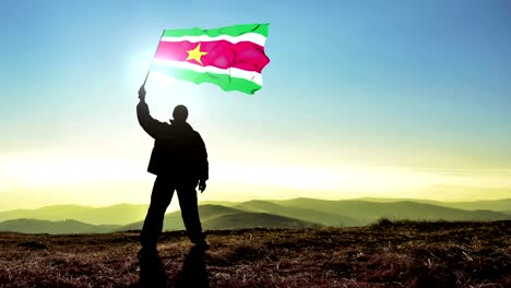 exitoso silueta hombre ganador agitando bandera de suriname en la parte superior del pico de la montaña, fondo de bucle cinemagraph