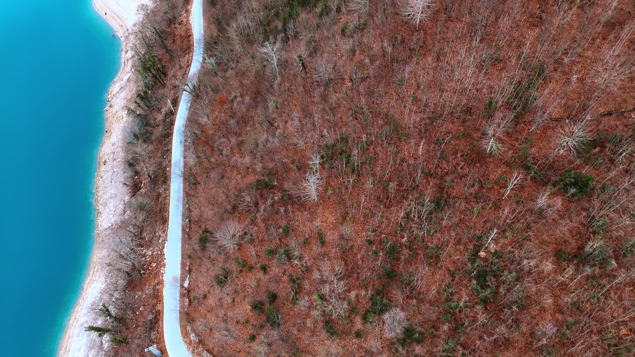 Top down aerial view of asphalt road by blue lake. Curving road separates turquoise water from a forest of bare trees with orange and brown leaves