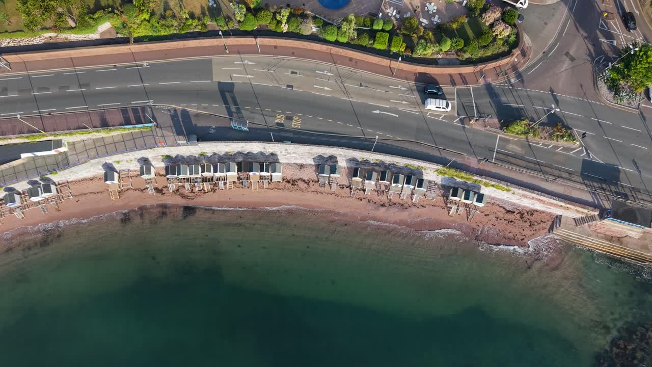 High aerial top-down drone shot of Torquay, UK. Blue sea and waves meet the beach, beach huts, wall, and main road with cars. Drone moves left, bending to follow the sweeping coastline
