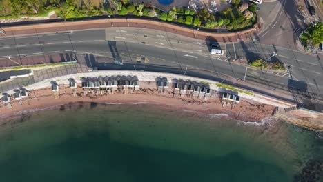 High aerial top-down drone shot of Torquay, UK. Blue sea and waves meet the beach, beach huts, wall, and main road with cars. Drone moves left, bending to follow the sweeping coastline
