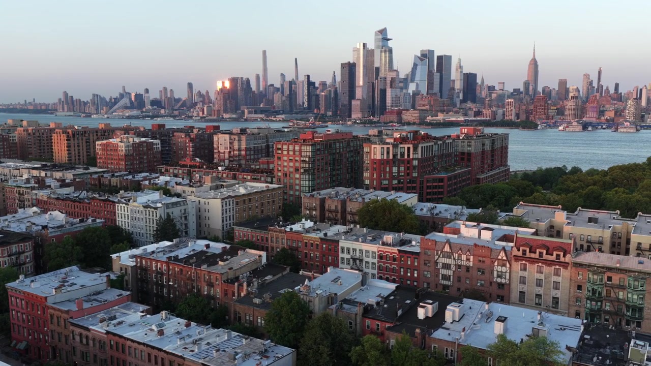 Teglrøde hustage fylder Hoboken-kvarteret, Manhattans skyline rejser sig bag Hudson River med Empire State Building, der gløder under en gylden solnedgang i New Jersey. Bredt droneoverblik.
