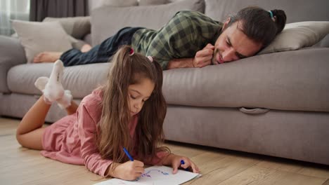 Un-Padre-Solitario,-Un-Hombre-Moreno-Con-Una-Camisa-A-Cuadros-Verde,-Se-Recuesta-En-El-Sofá-Y-Observa-A-Su-Pequeña-Hija-Con-Un-Vestido-Rosa-Dibujar-En-Una-Hoja-De-Papel-Blanca-Y-Acostarse-En-El-Suelo-En-Un-Apartamento-Moderno.