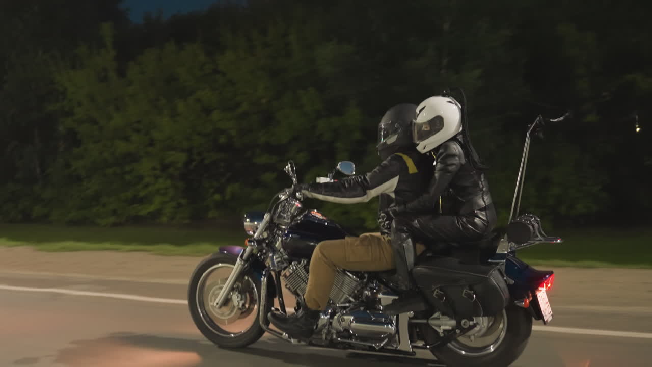 Side view of rescue team on motorcycle riding during night mission, wearing helmets, passing roadside electric pole while moving swiftly along asphalt road with green trees in background