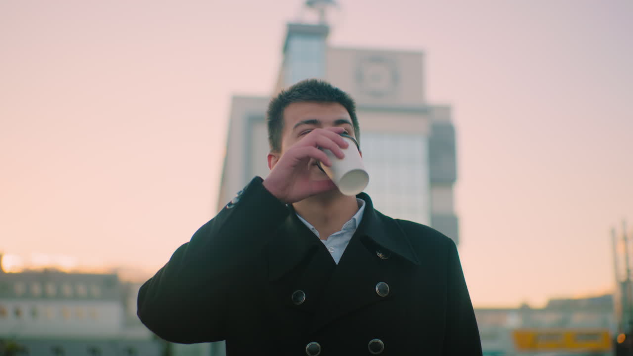 사업가 검은 슈트 야외에서 황혼에 자신감 있는 표정으로 커피를 마시고, 배경에는 현대적인 건물, 주차된 자동차 및 구조물이 있습니다.