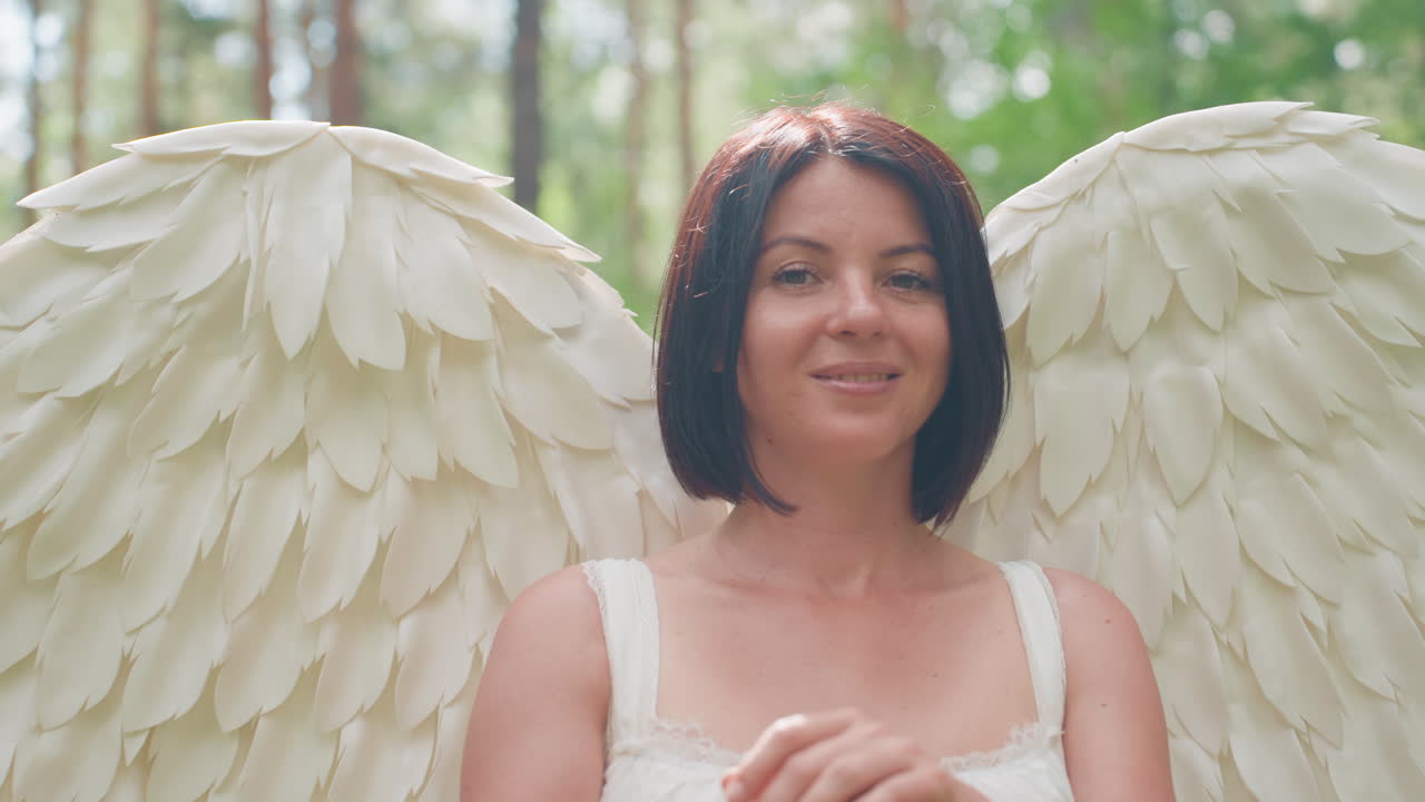 Angel woman dressed in white with large feather wings standing calmly in forest holding wooden staff under warm sunlight surrounded by lush green trees exuding serene presence grace