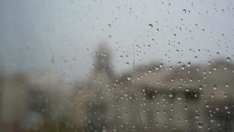 Una-Cámara-Lenta-De-Gotas-De-Lluvia-Vistas-A-Través-De-Una-Ventana-Durante-Fuertes-Lluvias,-Con-Un-Paisaje-Urbano-Al-Fondo