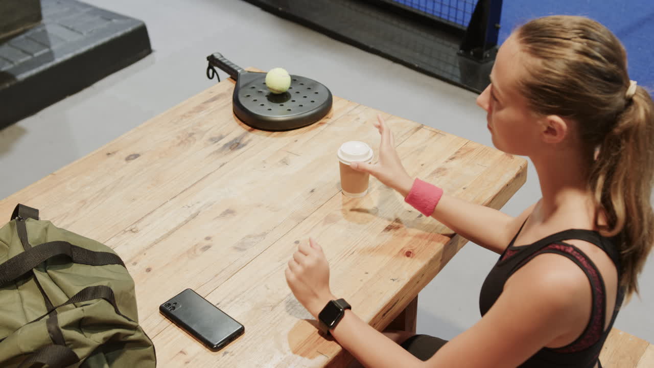 Diverse athletes relaxing after padel tennis match, enjoying coffee and conversation, on indoor cour