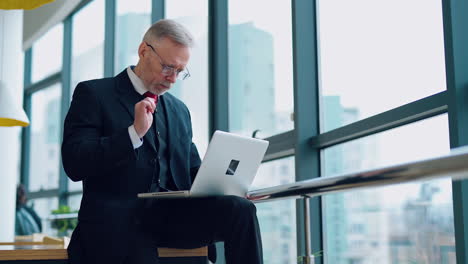 senior businessman in suit working near office window. elderly elegant man is typing on a laptop while sitting near the window. business concept.
