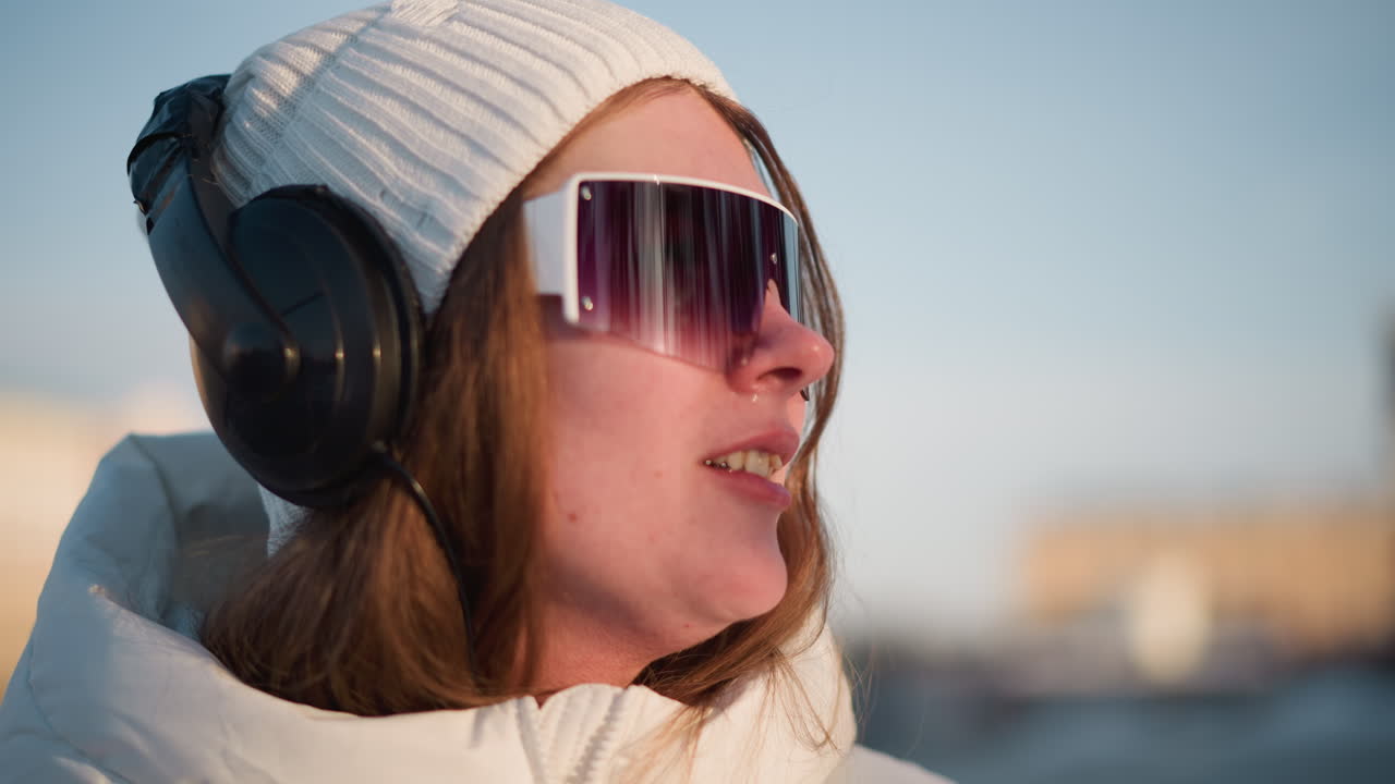 Close up portrait of young artist spinning outdoors in winter coat and beanie wearing sunglasses and headphones under warm sunlight head turn capturing motion against blurred scene with lens flare