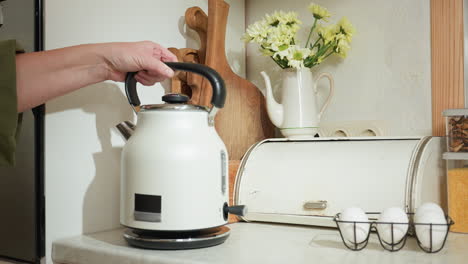 partial view of woman placing electric kettle on stove base in kitchen, switching it on as light indicator blinks, surrounded by rustic decor with eggs, cutting boards, and flower vase on counter