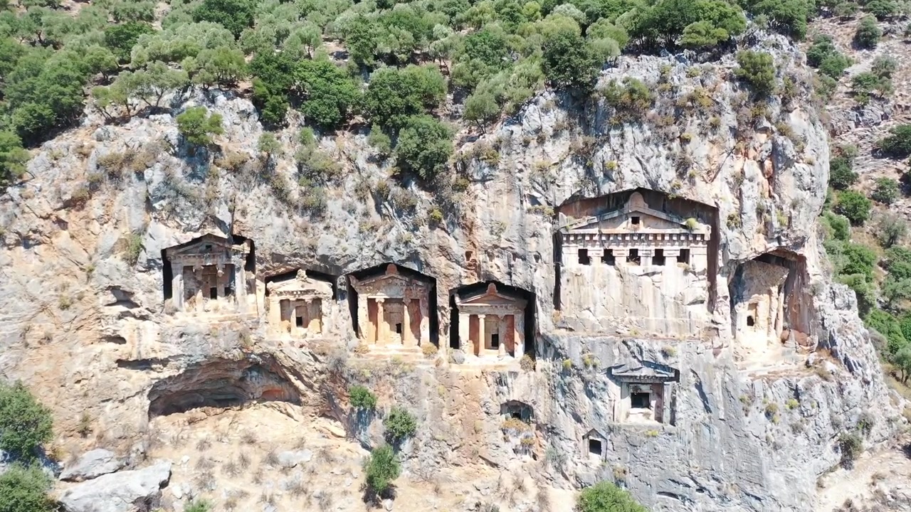 Güneşli bir günde, Türkiye'nin Muğla iline bağlı Dalyan'da, yeşil dağ yamaçlarıyla çevrili yüksek kaya yamaçlarına doğrudan oyulmuş antik Likya kaya mezarlarının havadan drone çekimi