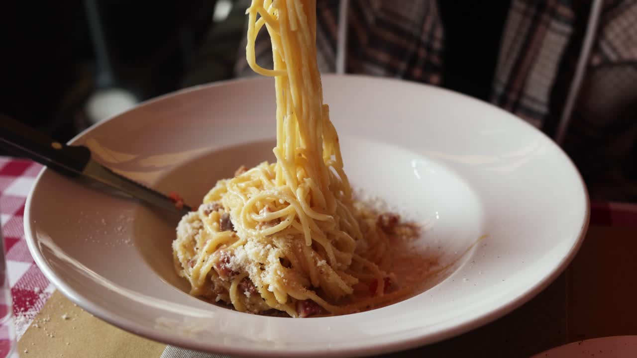 Mano che con la forchetta avvolge spaghetti alla carbonara cremosi, conditi con formaggio grattugiato e pancetta, in una ciotola bianca su una tovaglia a quadri rossi.