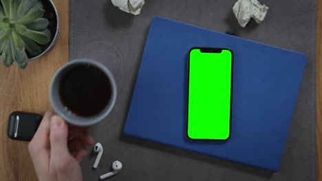 a man takes cup of coffee, standing on desktop next to mobile with green screen