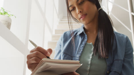 video of thoughtful biracial woman sitting on stairs and making notes