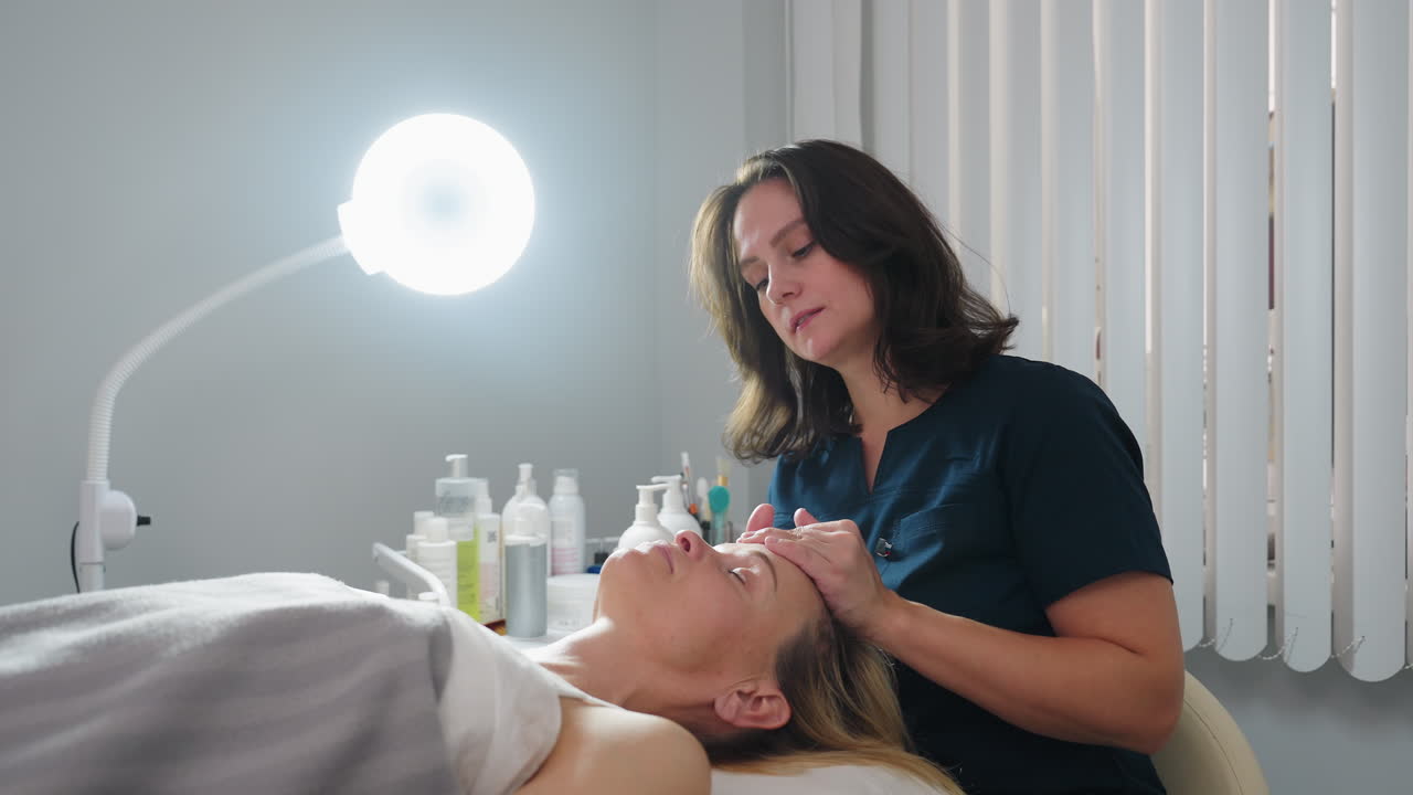 Facial practitioner massaging face from cheeks to forehead with gentle fingertip glides while client closes eyes in serene spa environment with ring light and skincare products visible beside bed