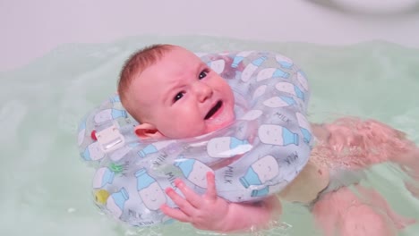 sad infant baby boy crying while swimming in the bathtub with an inflatable ring on his neck. four month old child, close-up