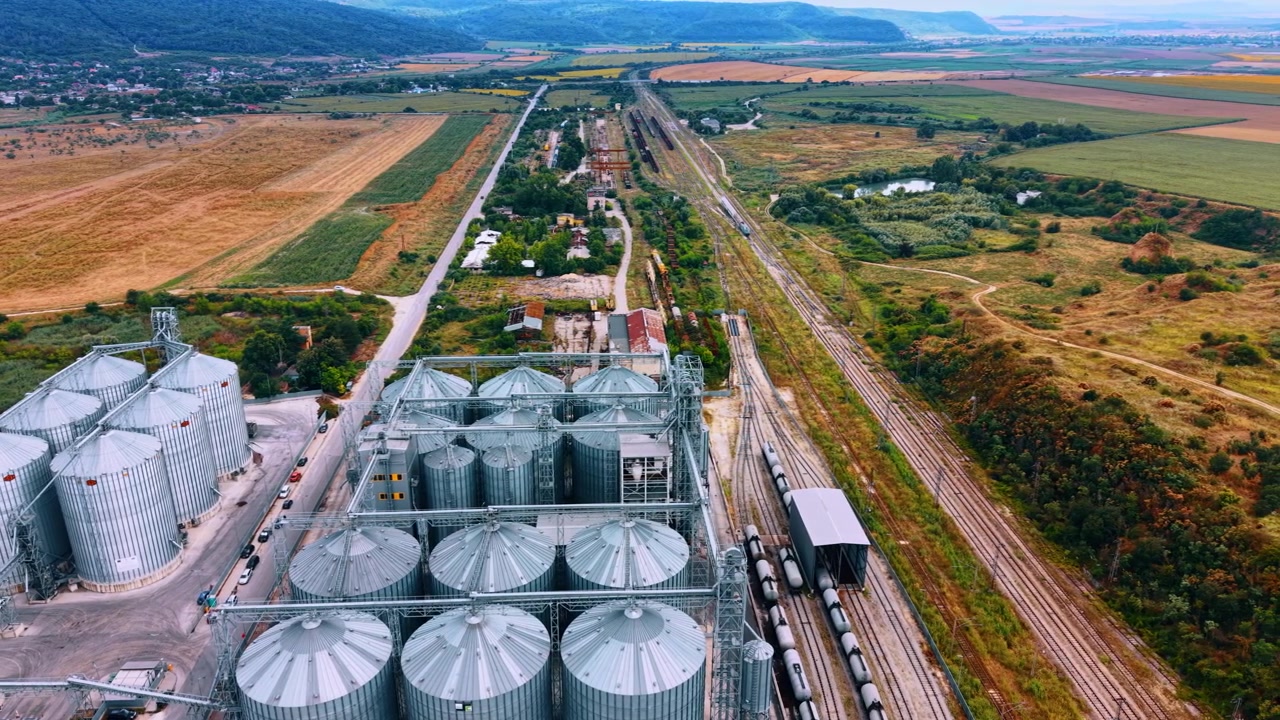 Getreideterminal neben einer Bahnstrecke in einer Agrarlandschaft. Weitwinkel-Drohnenansicht von Metallsilos, Fördertürmen, Kesselwagen, mehreren Gleisen, Feldern, Straßen und entfernten Hügeln.