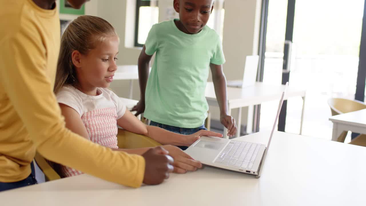 Diverse kids at desk girl pointing to silver laptop boy yellow reaching trackpad watching for work