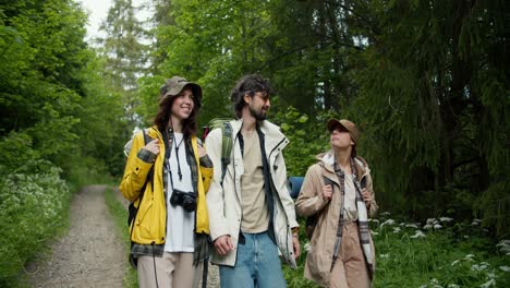 a guy and two girls in special hiking clothes are walking along a path along a green forest. chatting while walking through the woods