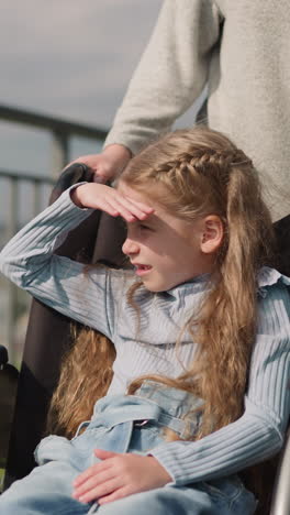 little girl with cerebral palsy enjoys city view covering eyes from bright sunlight. woman walks on street pushing wheelchair with daughter closeup