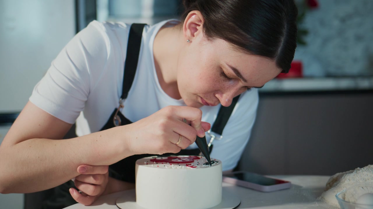 Ung kvinne dekorerer forsiktig en liten kake med glasur. Hun fokuserer intenst på arbeidet sitt, viser tålmodighet og dyktighet. Perfekt for matblogger, bakerireklamer eller kulinariske veiledninger.