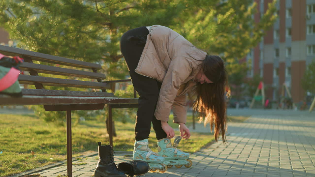 A young woman wearing a peach jacket and black trousers is bent over, fastening the straps of a rollerblade on her feet. The setting is an outdoor park with a bench nearby