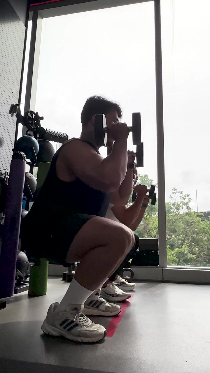 Dua pria melakukan squat dumbbell di gym terang dengan jendela besar dan cahaya alami