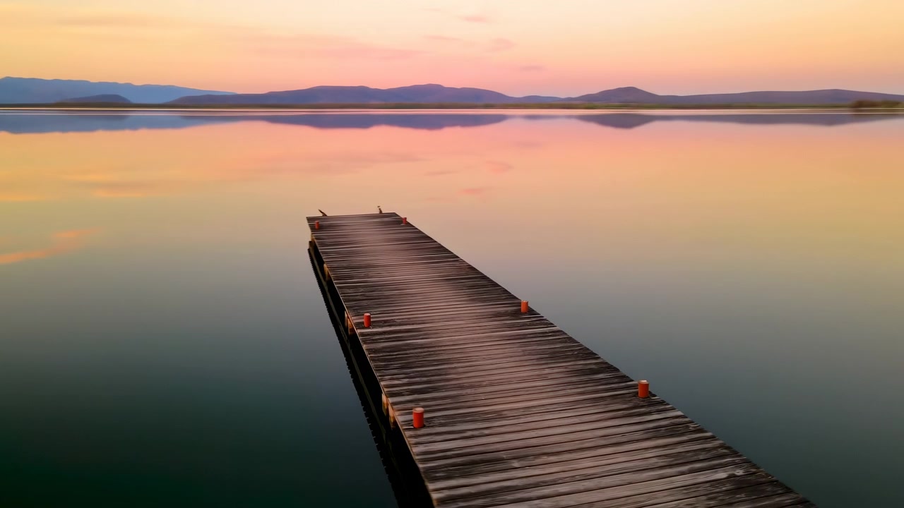 Un pontile di legno stagionato, centrato nell'inquadratura, si estende verso un lago calmo al tramonto, con un cielo rosa pastello e arancione che si riflette sull'acqua immobile e una bassa distanza