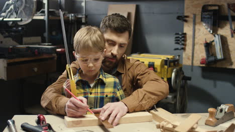 caucasian carpenter man teaching his little son to work with hardwood and sawing timber in workshop