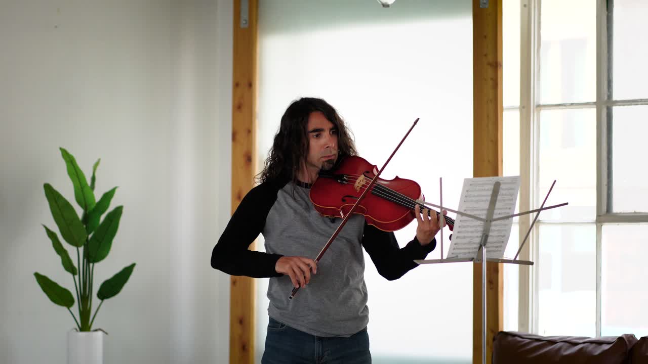 Inquadratura media di un musicista maschio ispanico con capelli lunghi, in maglia a maniche lunghe grigia e nera, che esegue una canzone da uno spartito su una viola rossa
