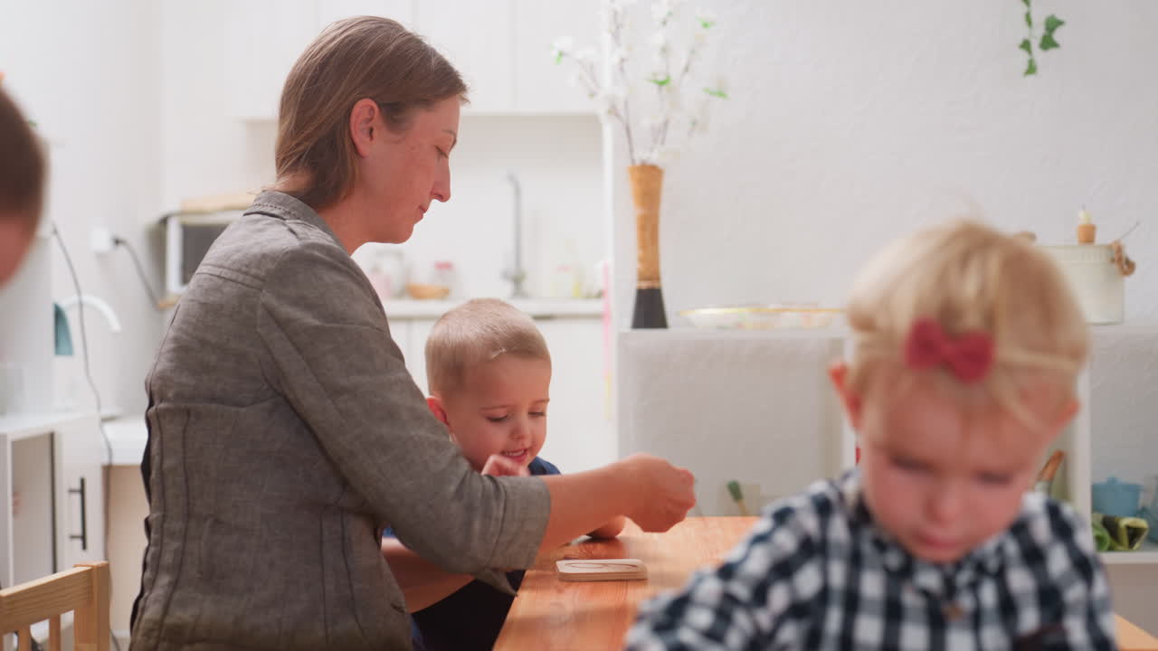 Un profesor ayuda a un niño pequeño con un puzle de madera durante una clase preescolar en un aula luminosa, guiándole con las manos sobre la mesa mientras una niña pequeña en primer plano se mueve de forma borrosa, un momento cálido y enriquecedor de educación en interiores