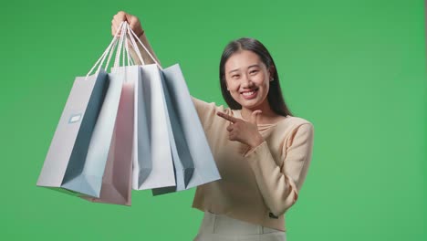 An Asian Shopping Woman Holding Shopping Bags Up Before Pointing And Smiling To Camera While Standing In Front Of Green Screen