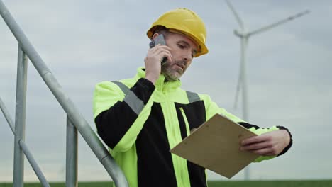 hombre profesional caucásico de pie en el campo de la turbina eólica y hablando por teléfono móvil.