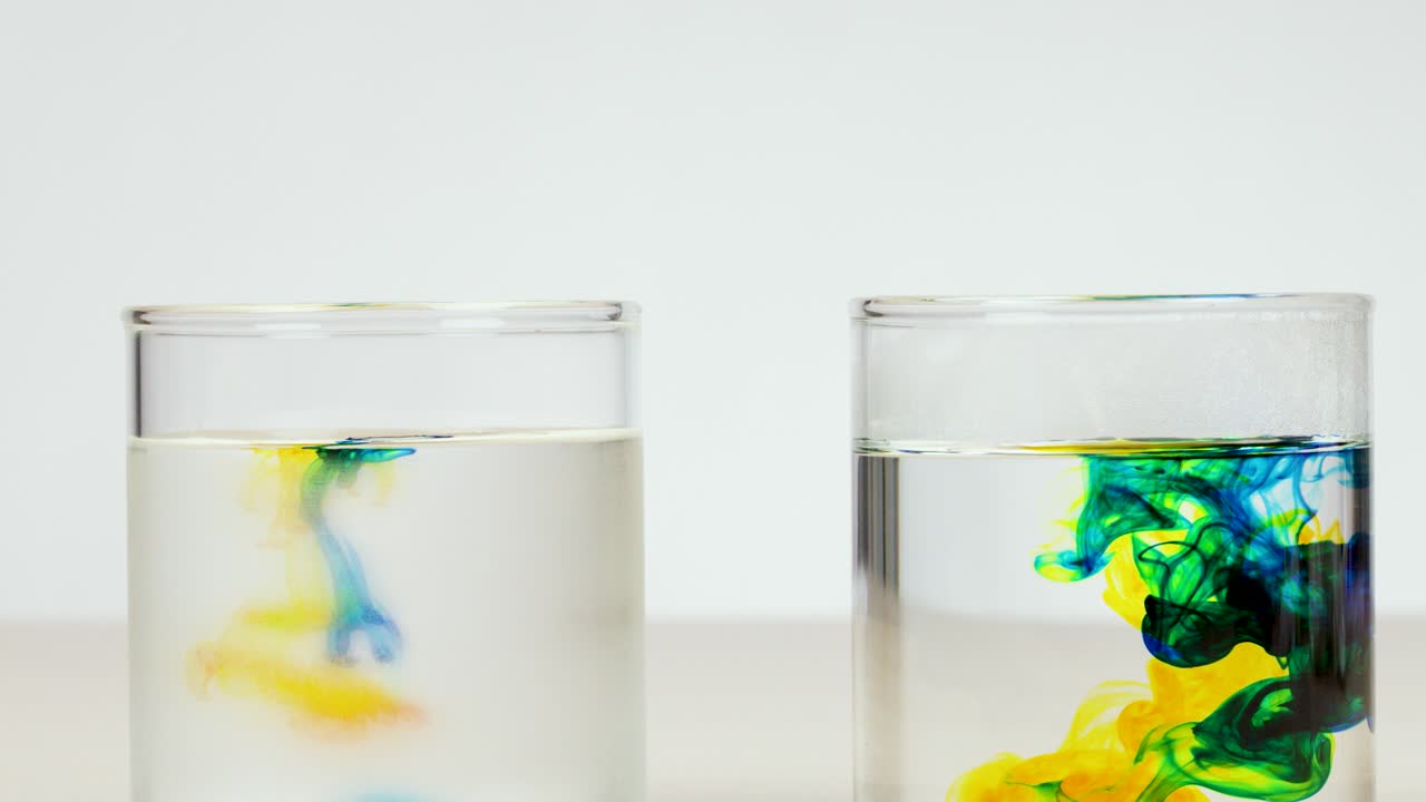 Colored drops disperse in clear water, demonstrating diffusion in a bright, minimal studio setting