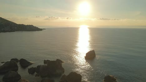 aerial shot of a scenic sunset in the mediterranean sea, with big rocks in the water and the sun reflecting on the surface of the sea, malta