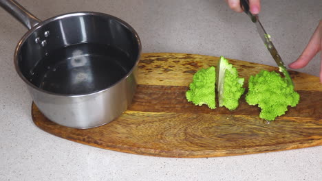 female cook cutting fresh baby romanesco broccoli - cauliflower with a sharp knife on a wooden chopping board, adding the chopped pieces to a pan of water