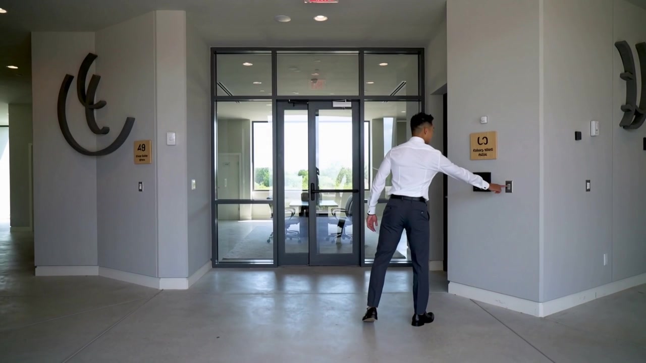 Hombre caminando con camisa blanca y pantalones oscuros acercándose a las puertas de cristal de la oficina, presionando el panel y entrando
