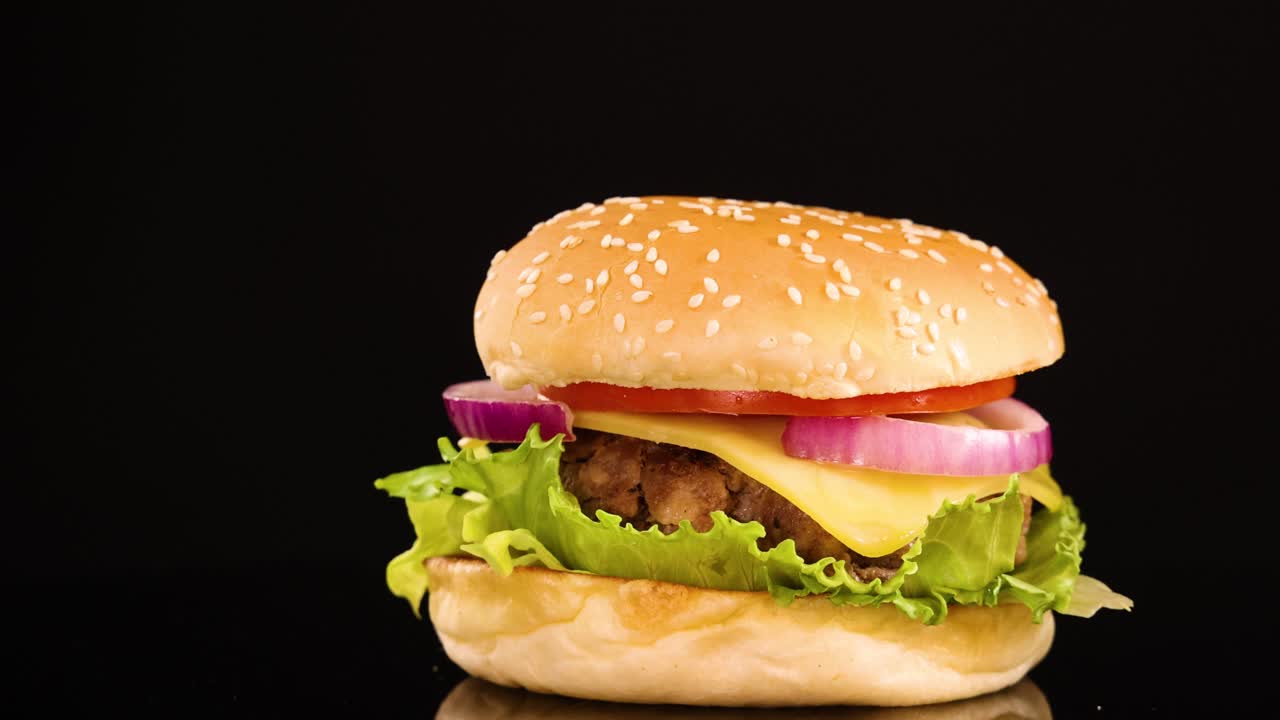A classic beef burger with lettuce, tomato, onion, and sesame bun rotates smoothly against a black backdrop under bright, even studio lighting