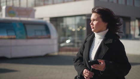 lady walking outdoors in a black coat and white turtleneck, holding her phone and glancing around with a thoughtful expression, background features modern urban buildings