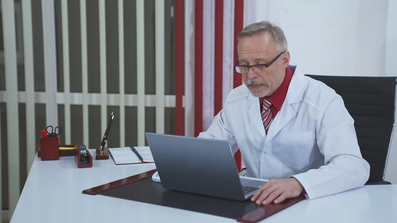 Older doctor making video call on laptop. Senior male physician talking to patient while looking at pc screen communicating by webcam in web chat. Telemedicine concept.