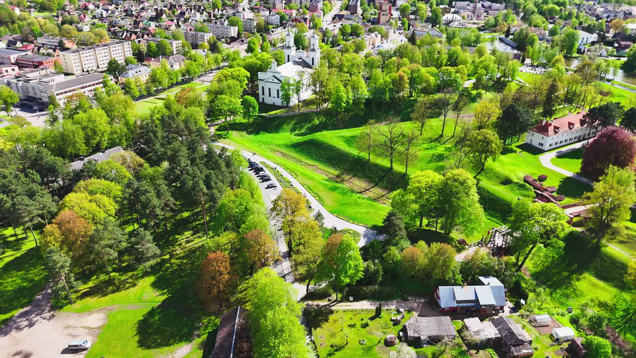 A vibrant European town is surrounded by blooming spring trees, winding park paths, and lush open spaces, captured from above on a sunny day.