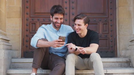 Una-Pareja-Gay-Masculina-Sentada-Al-Aire-Libre-En-Las-Escaleras-Del-Edificio-Mirando-El-Teléfono-Móvil